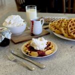 Buttermilk waffles with berry sauce and whipped cream. Glass of milk, plate of waffles, and jar of berry sauce.